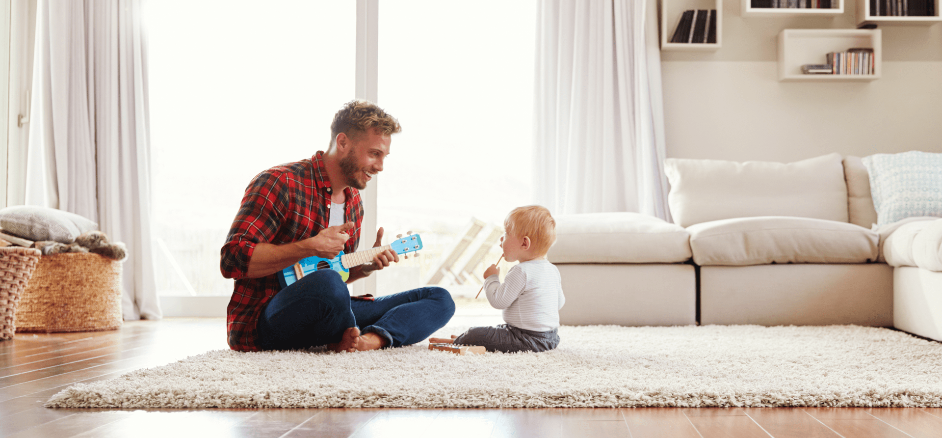 Dad smiling as he plays with young toddler’s toy guitar, toddler looks up at him in a light and airy room