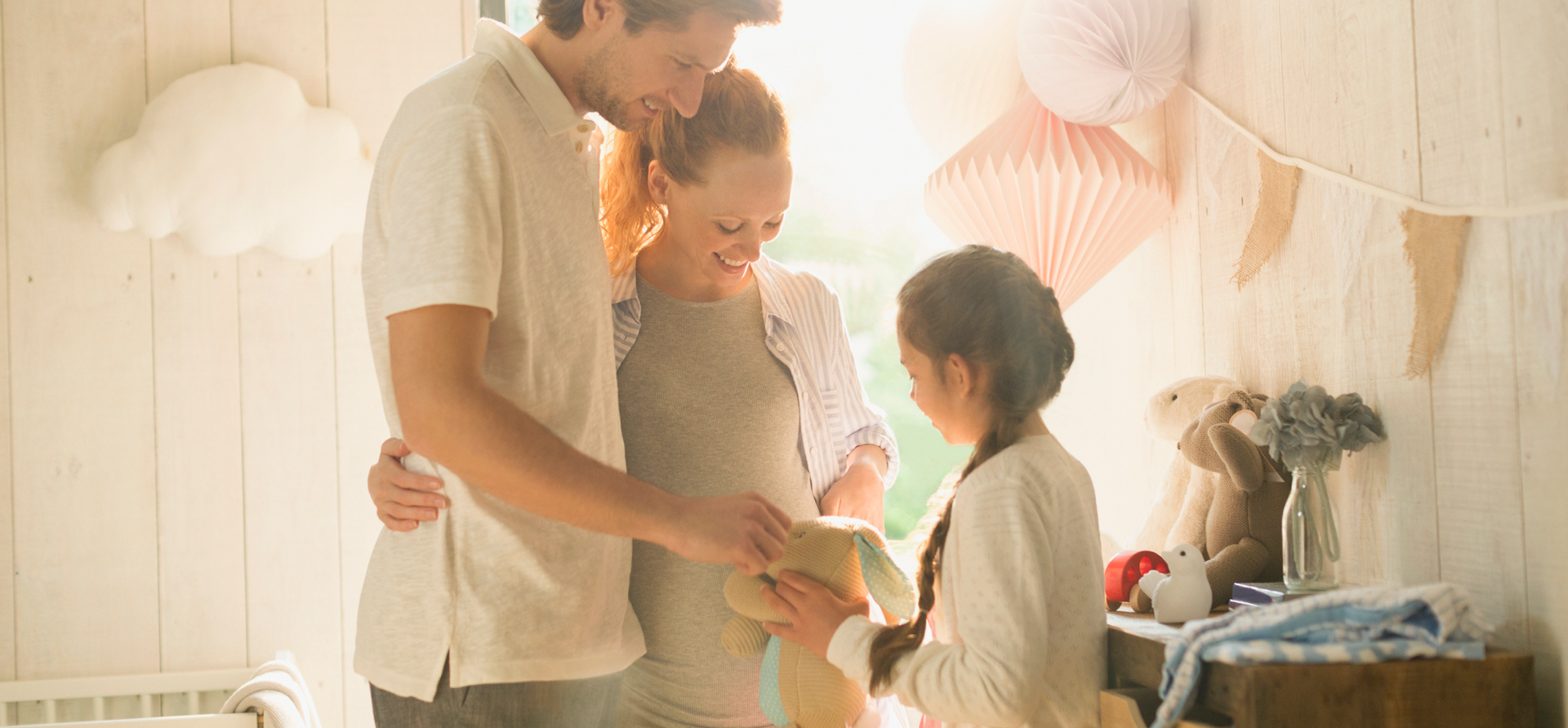 A pregnant woman, her young man partner, and a young girl all smiling and admiring a bunny toy in a room with sunlight shining through the window.