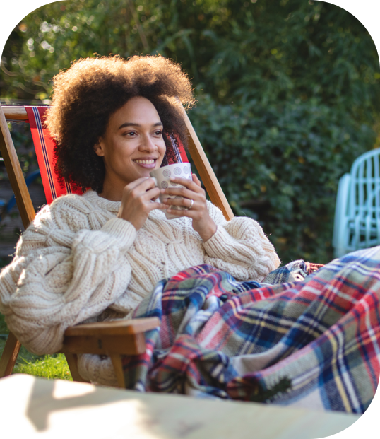 Woman relaxing outside on the patio chair with a blanket wearking a sweater and drinking a refreshment.